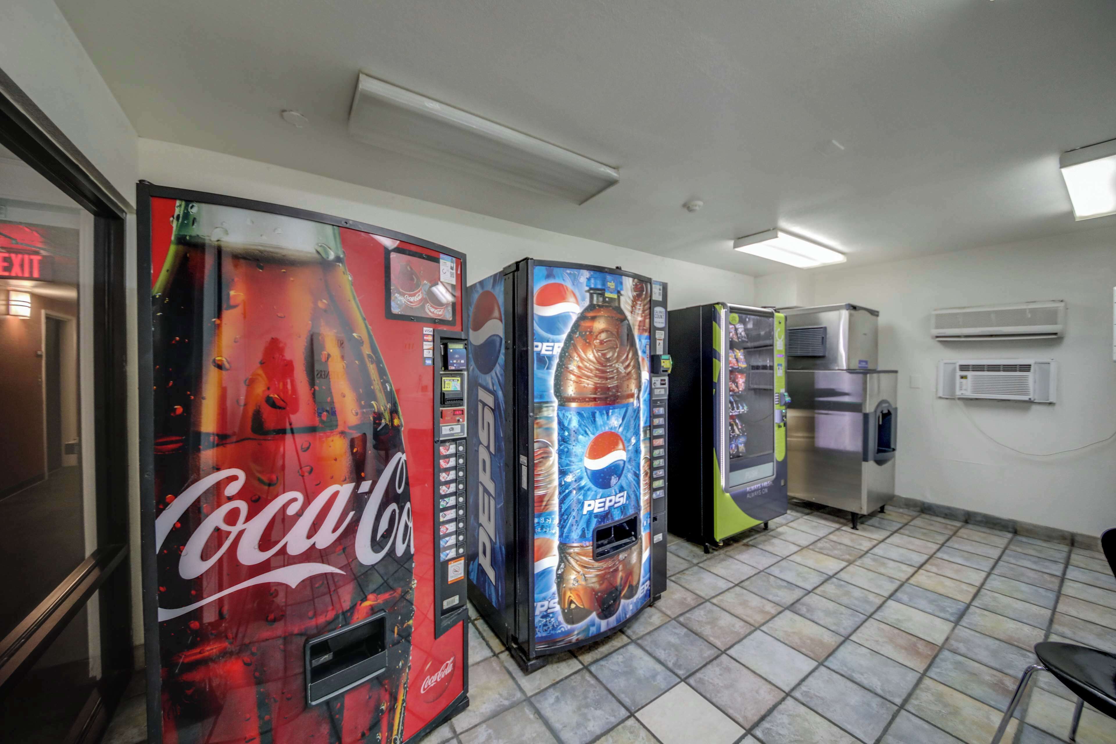 Dans une pièce dont le sol est recouvert de carreaux, on trouve des distributeurs automatiques aux couleurs de Coca-Cola et de Pepsi, un réfrigérateur et un appareil de climatisation.