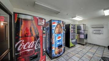 Dans une pièce dont le sol est recouvert de carreaux, on trouve des distributeurs automatiques aux couleurs de Coca-Cola et de Pepsi, un réfrigérateur et un appareil de climatisation.