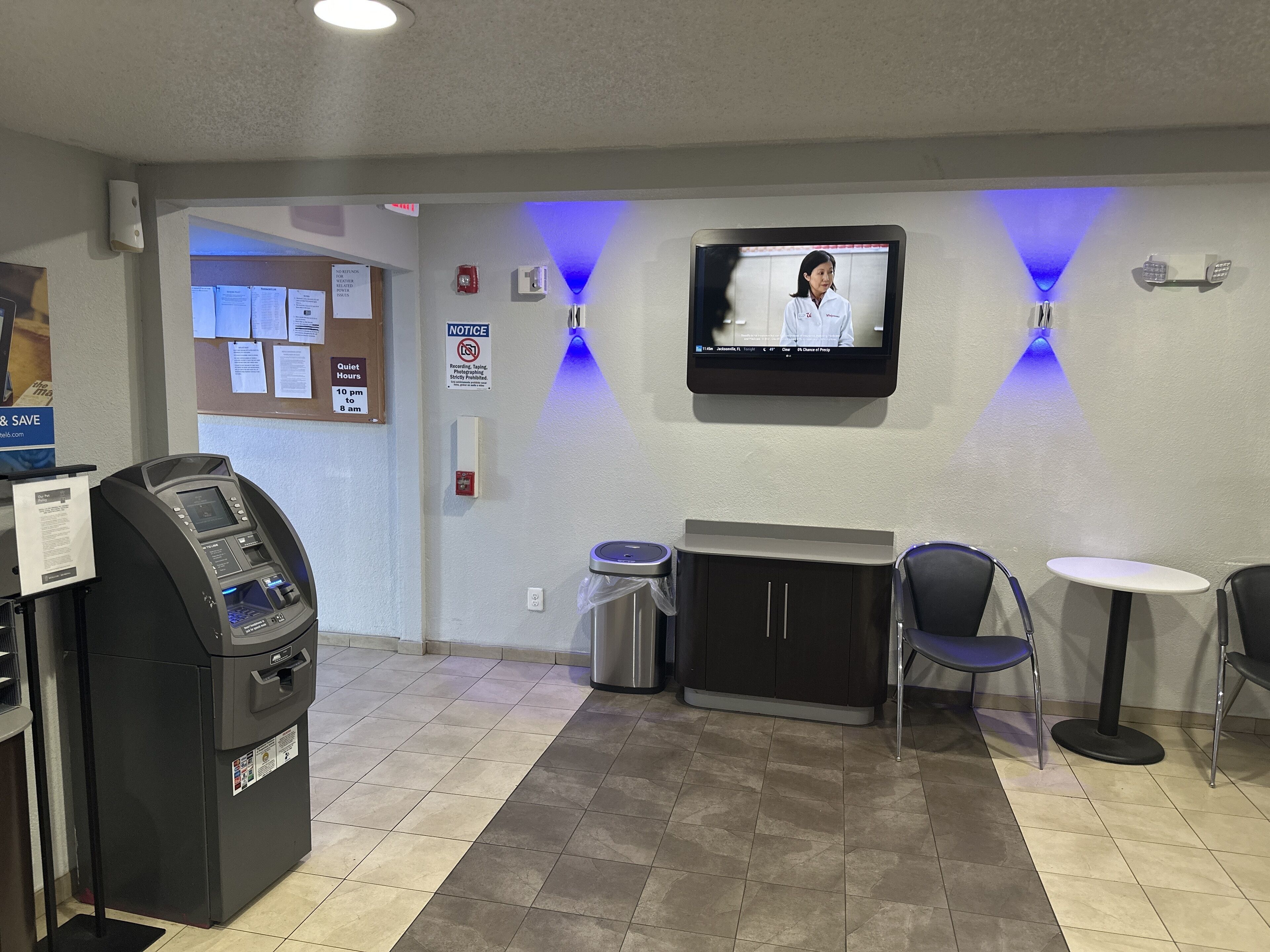 A lobby area with a self-service kiosk, a television mounted on the wall, a notice board, and a seating area with chairs and a small table.