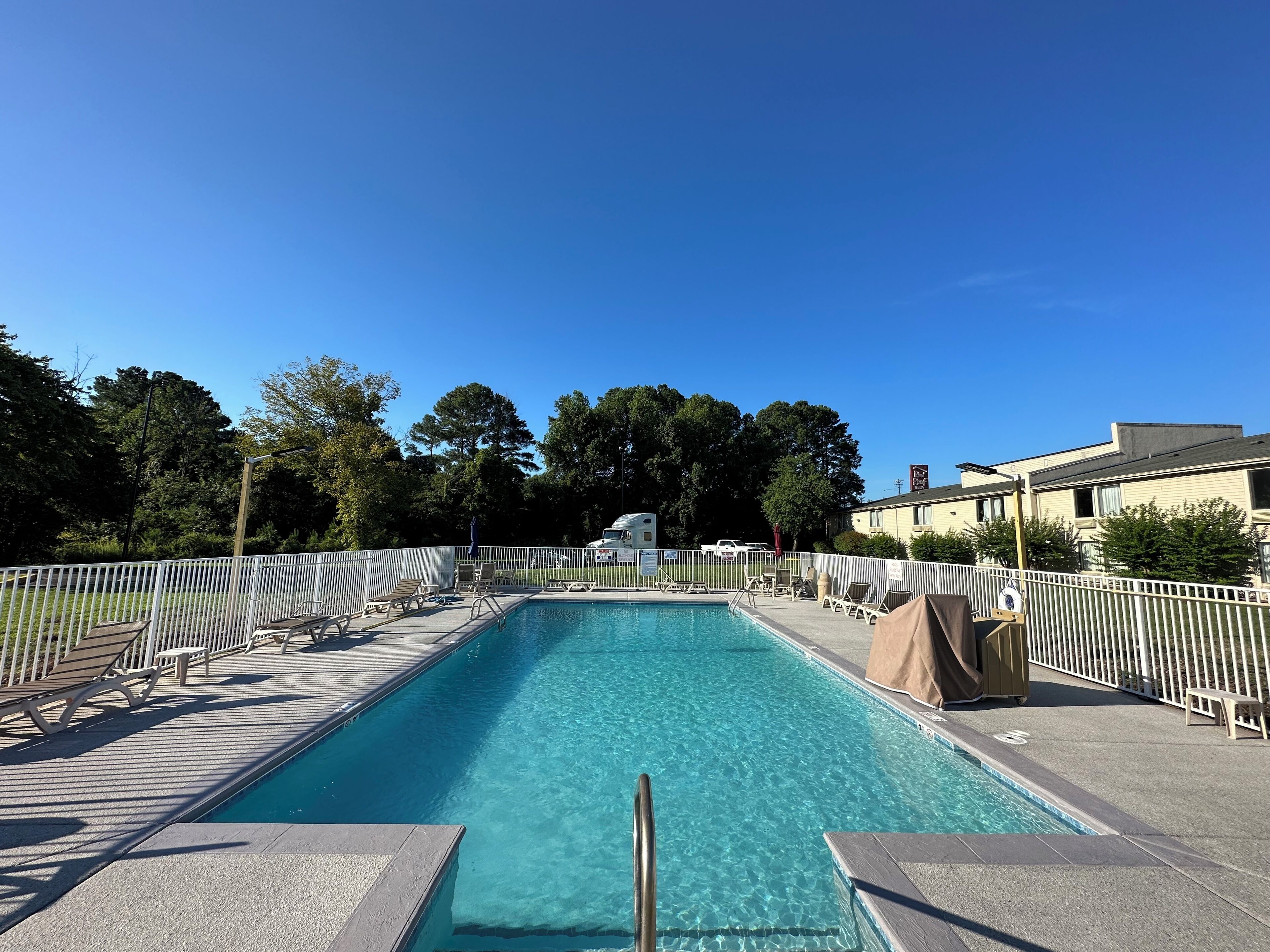 A rectangular swimming pool with a white fence surrounding it, clear blue water, and several lounge chairs along the sides.