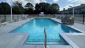 Ein rechteckiger Swimmingpool mit Metallhandlauf in der Mitte, umgeben von Liegestühlen und einem eingezäunten Bereich.