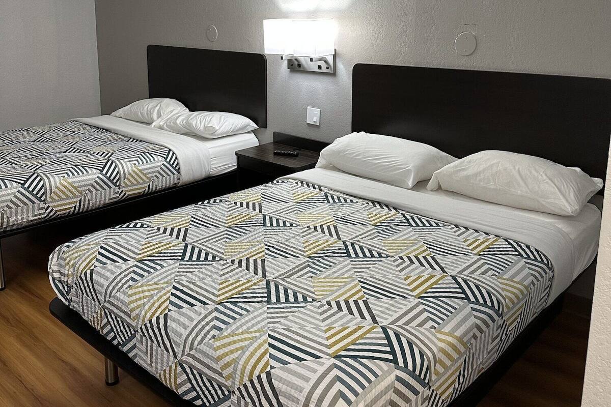 A hotel room with two single beds, a nightstand, a wall-mounted light, and a plain ceiling.