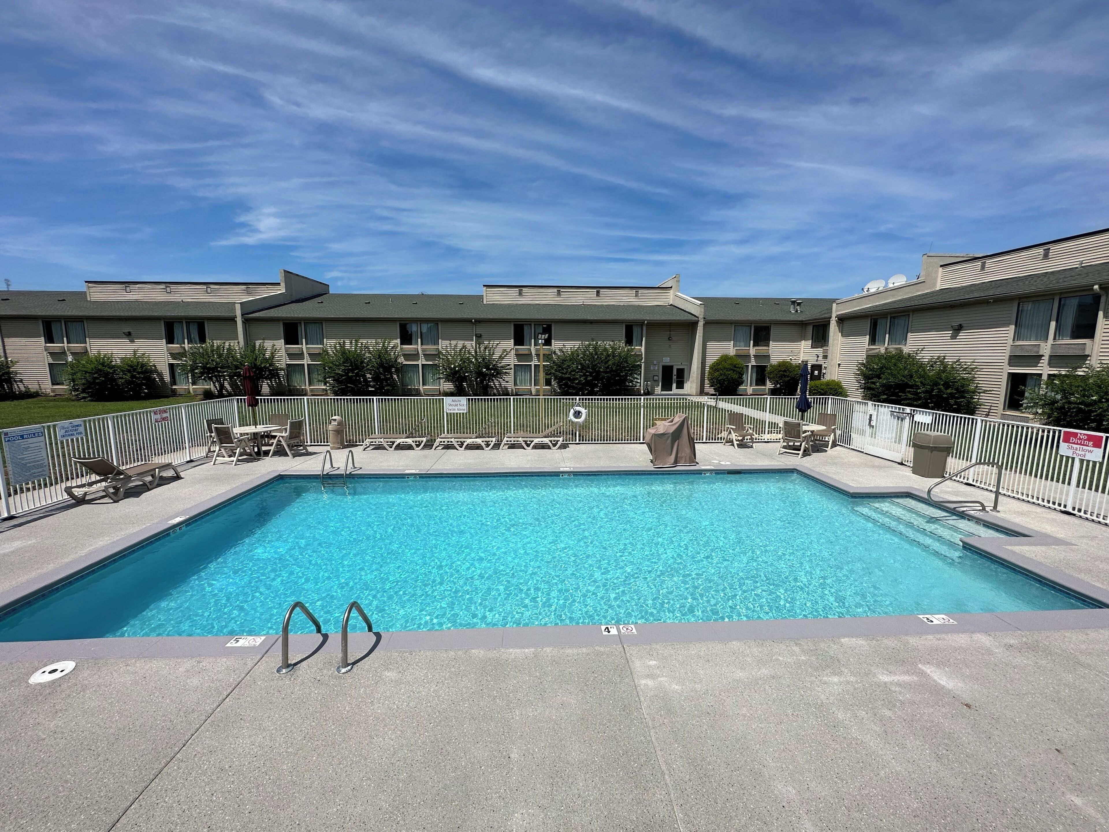 A rectangular swimming pool with clear blue water, surrounded by a white fence, and adjacent to a row of beige apartment buildings.
