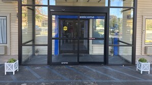 Ein moderner Gebäudeeingang mit Glastür, einem 'welcome'-Schild und einem 'caution stand back'-Schild.