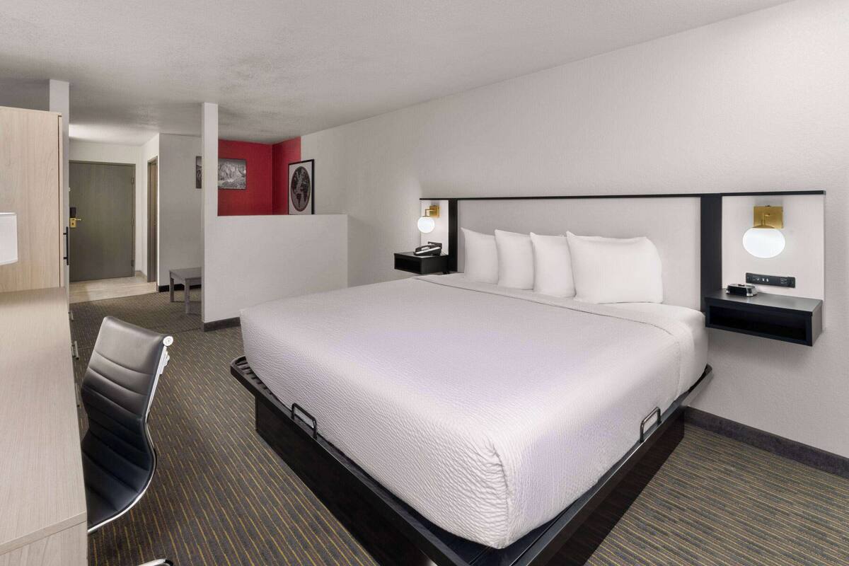 Ein Hotelzimmer mit einem großen Bett, einem Schreibtisch und einem Stuhl. Ein Nachttisch mit Lampe und kleiner Tisch sind vorhanden. Der Raum verfügt über eine rote Akzentwand und eine Tür zu einem weiteren Zimmer.