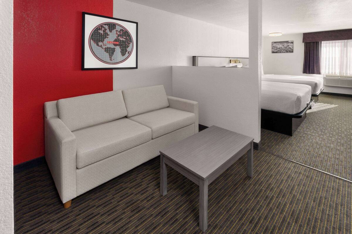 Ein Hotelzimmer mit grauem Sofa, einem kleinen Tisch, zwei Betten, einer gerahmten Weltkarte und einem Vorhangfenster.
