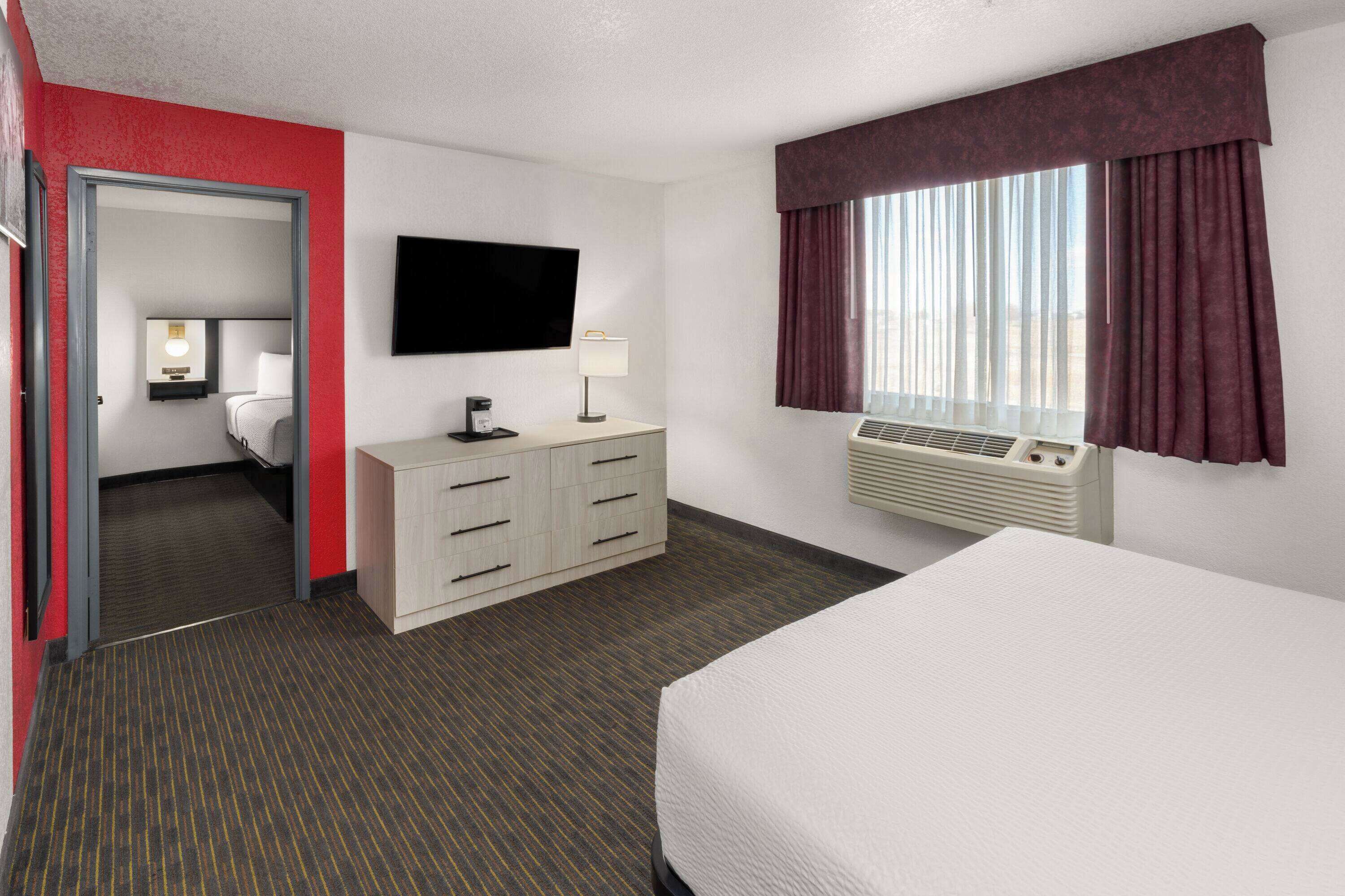 A hotel room with a bed, a desk, a television, a window with curtains, and a door leading to another room.