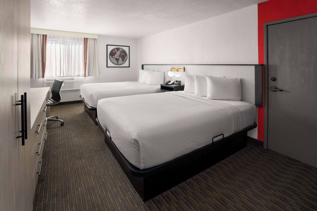 Ein Hotelzimmer mit zwei Betten, einem Schreibtisch, einem Stuhl, einer Schreibtischlampe und einem gerahmten Bild an der Wand.