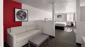 A hotel room with a white sofa, a small table, a bed, a window with curtains, and a framed world map on the wall. - SYLO Hotel Denver Airport, a Ramada by Wyndham (Aurora)