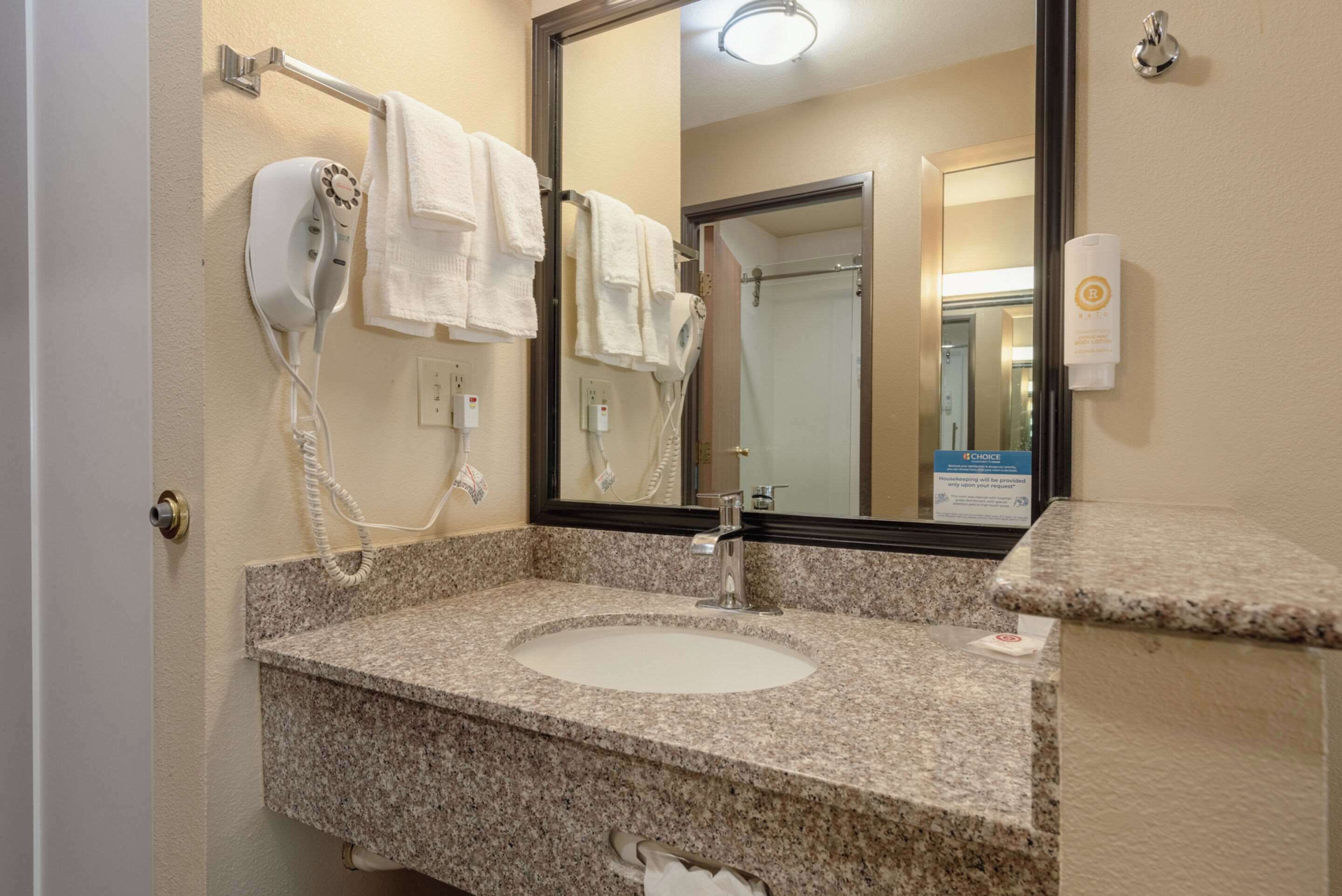 A bathroom with a granite sink, a towel rack with multiple white towels, a hairdryer, and a mirror reflecting a shower area.