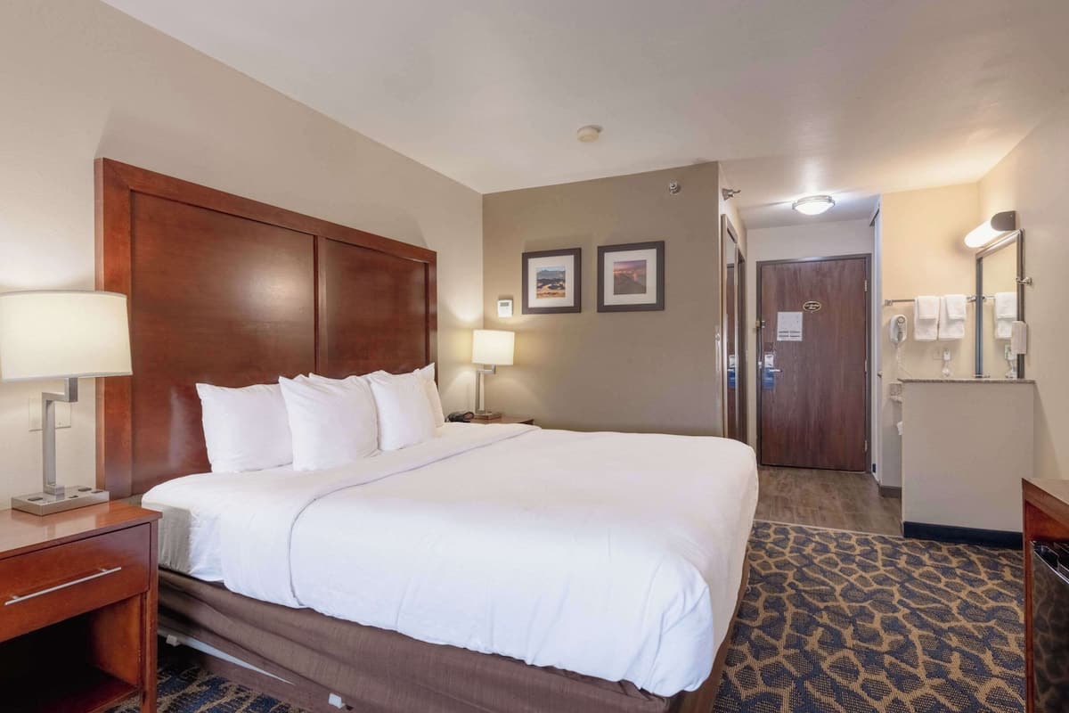 A hotel room with a large bed, a wooden headboard, a nightstand with a lamp, a bathroom with a mirror, and a patterned carpet.