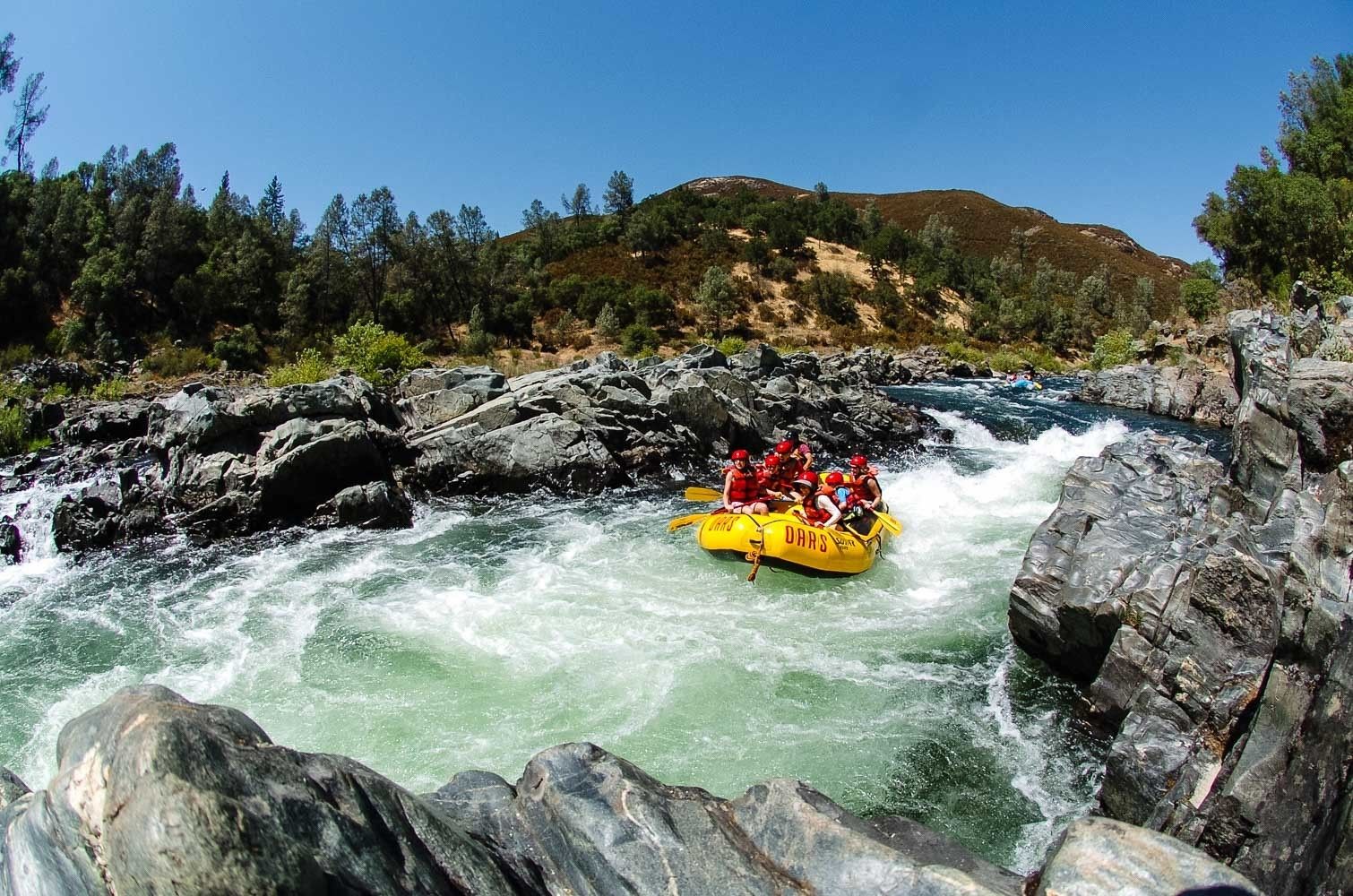 A group of people in a yellow raft navigating a river surrounded by rocky terrain and trees.