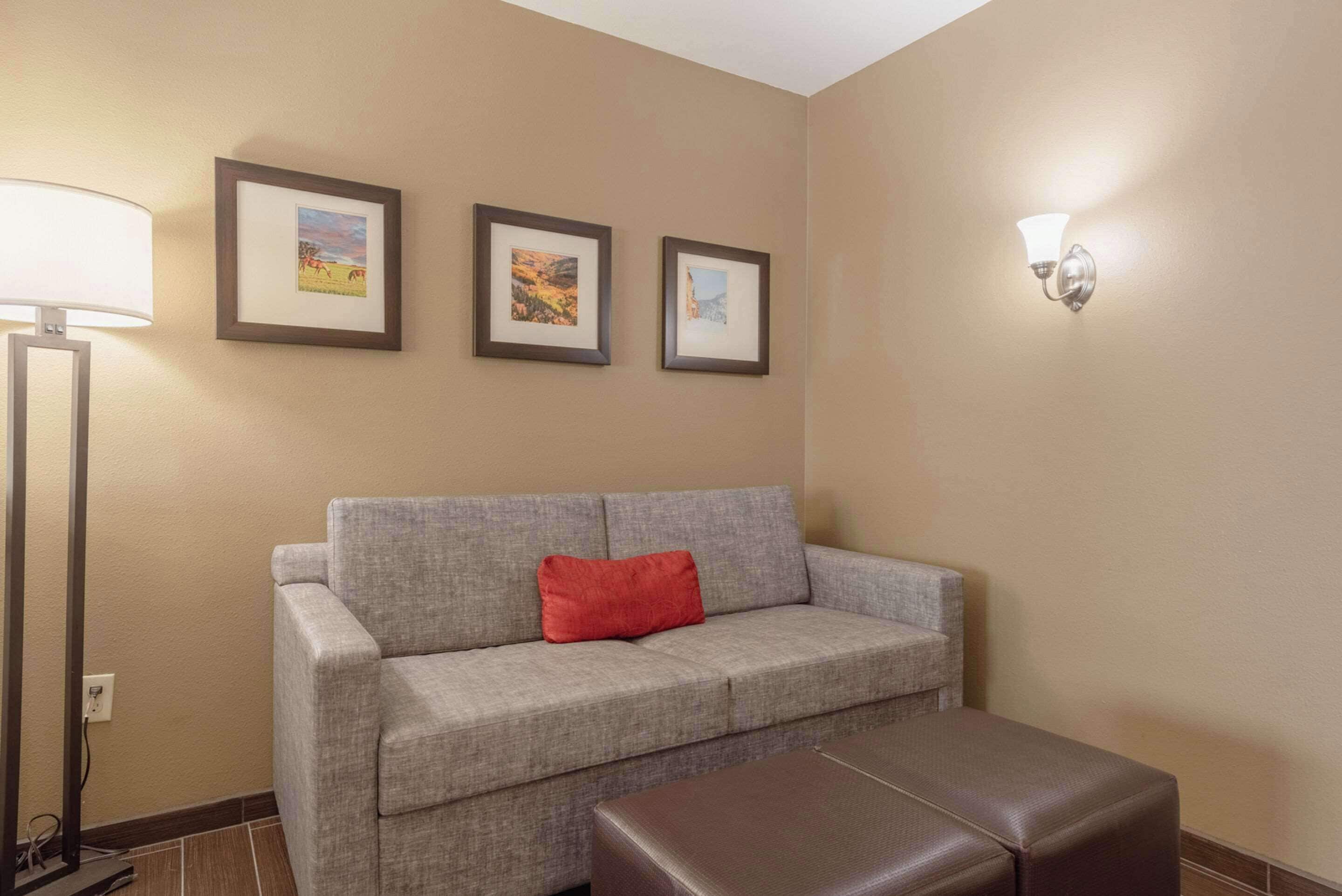 Una habitación en tonos neutros con un sofá gris, un cojín rojo, un reposapiés marrón y una lámpara de pie. Hay tres cuadros en la pared.