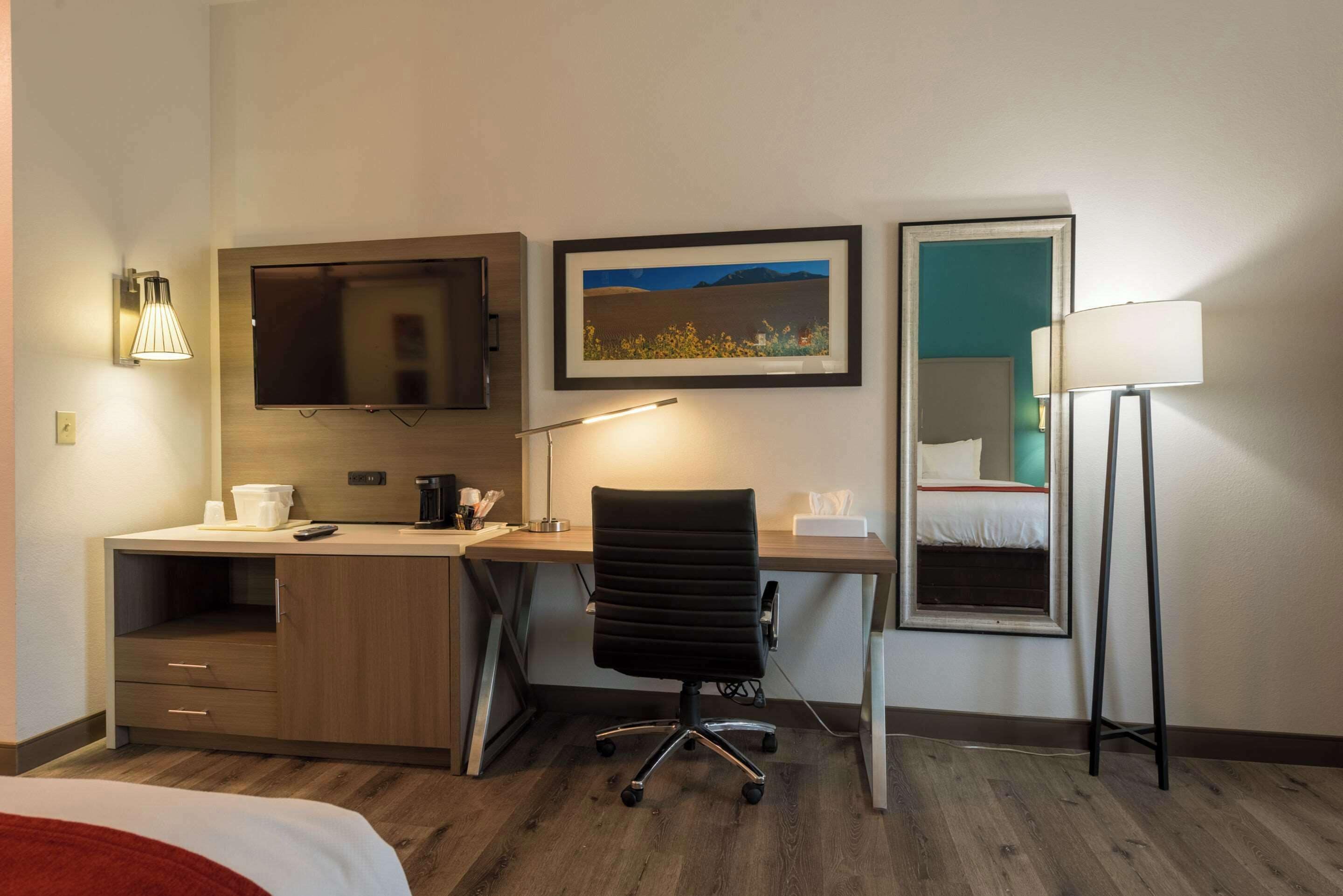 A hotel room with a wooden desk, a black office chair, a flat-screen TV, a framed landscape picture, a bedside lamp, and a bed with a red blanket.