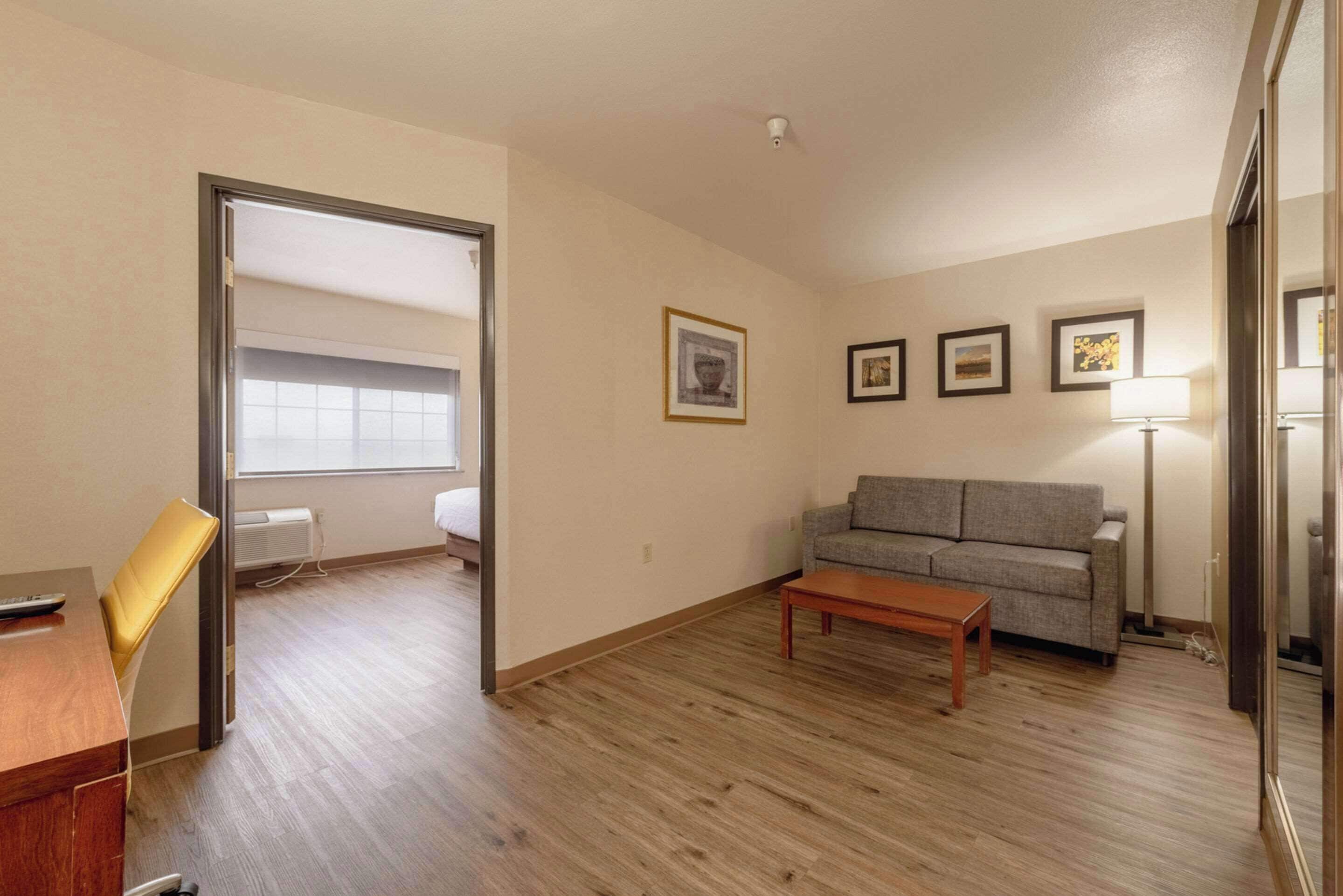 Amplia sala de estar con piso de madera, un sofá, una mesa de centro y un sillón. Hay una puerta que conduce a otra habitación y una ventana con persianas.