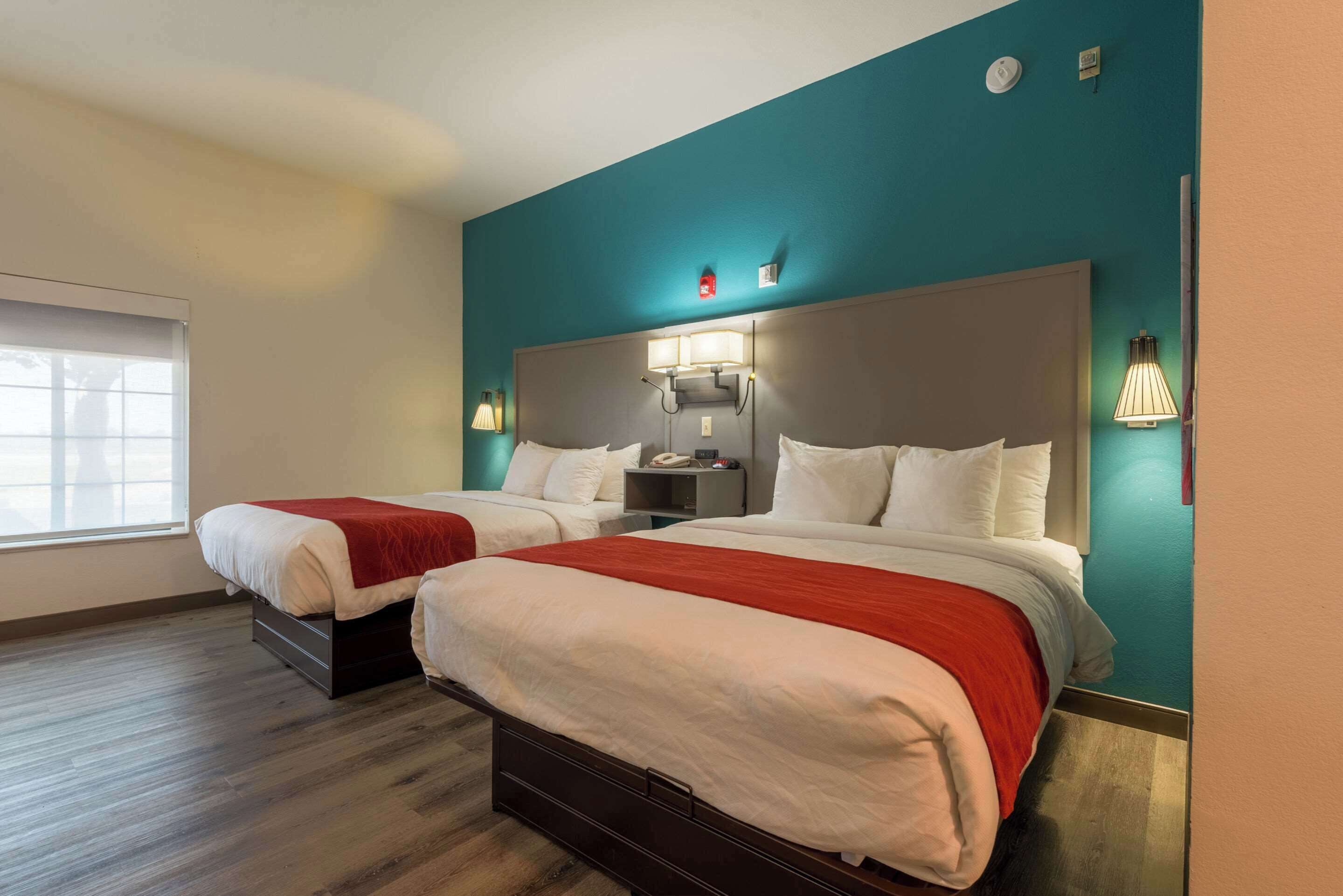 A hotel room with two beds, a teal accent wall, and a window with blinds.