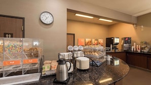 A hotel breakfast area with a coffee station, cereal, pastries, and a clock on the wall.