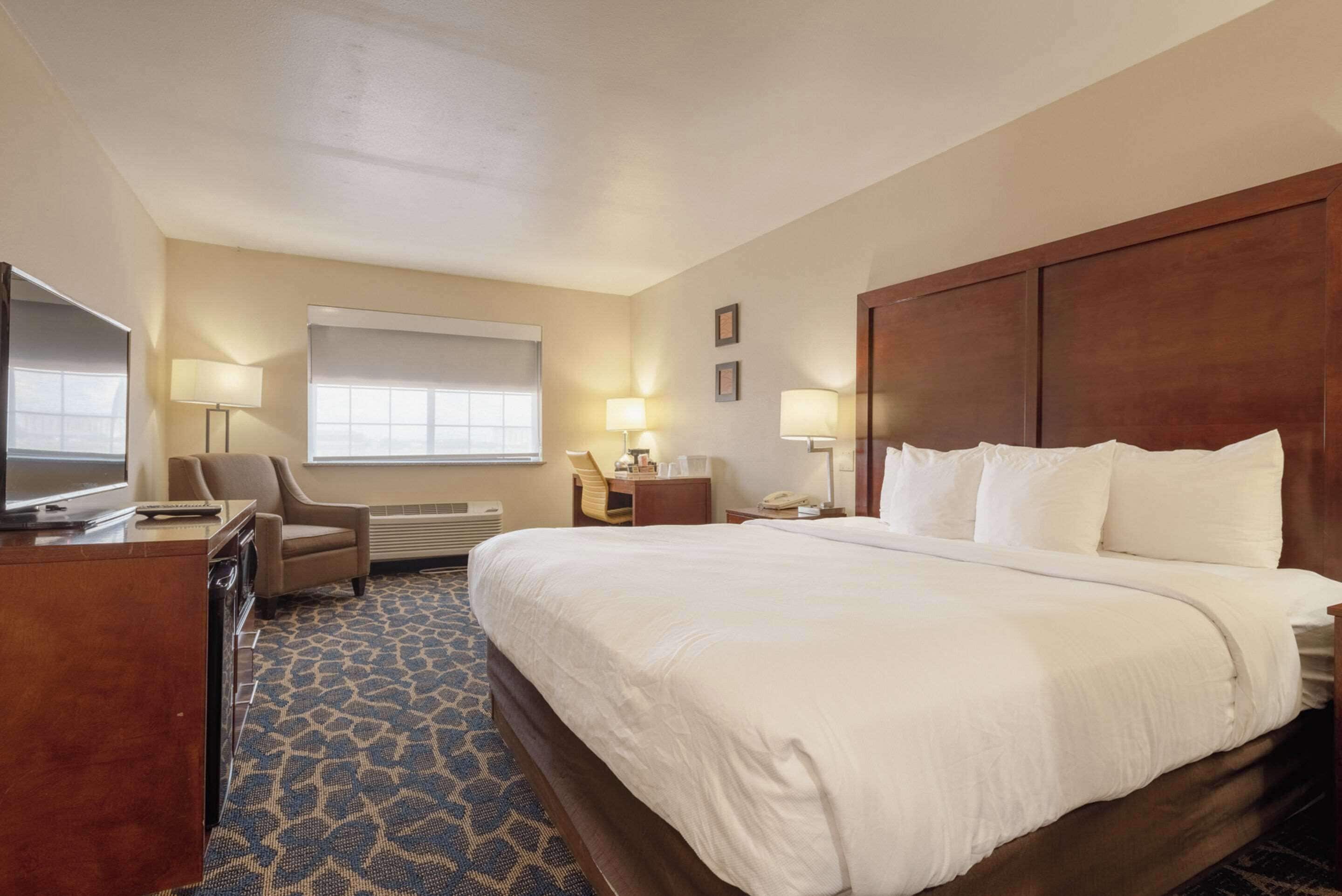 A hotel room with a large bed, a desk with a television, a chair, a desk lamp, and a window with blinds.