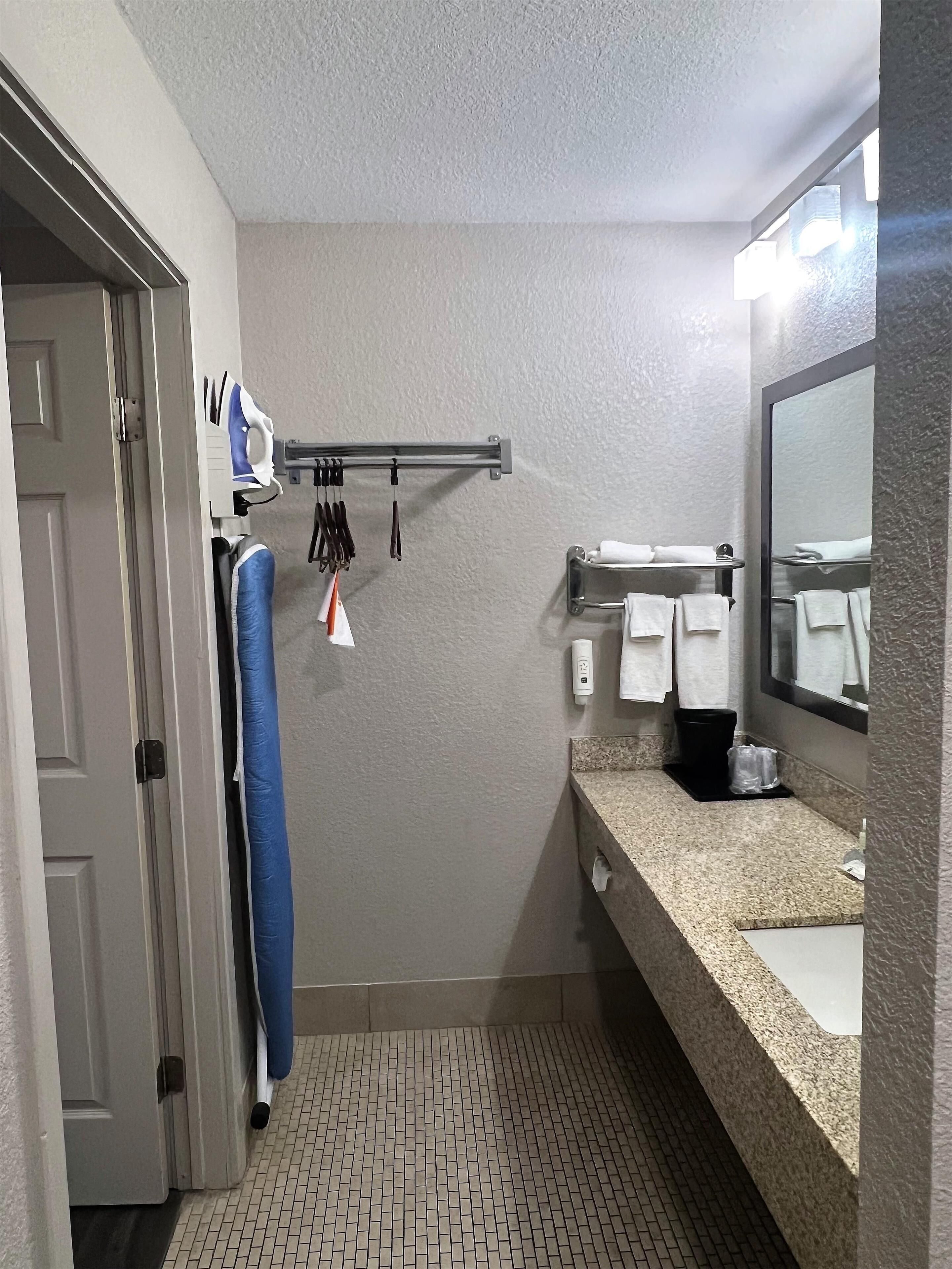 A bathroom with a tiled floor, a sink with a granite countertop, a towel rack with white towels, a mirror, and a door with a towel hanging on it.