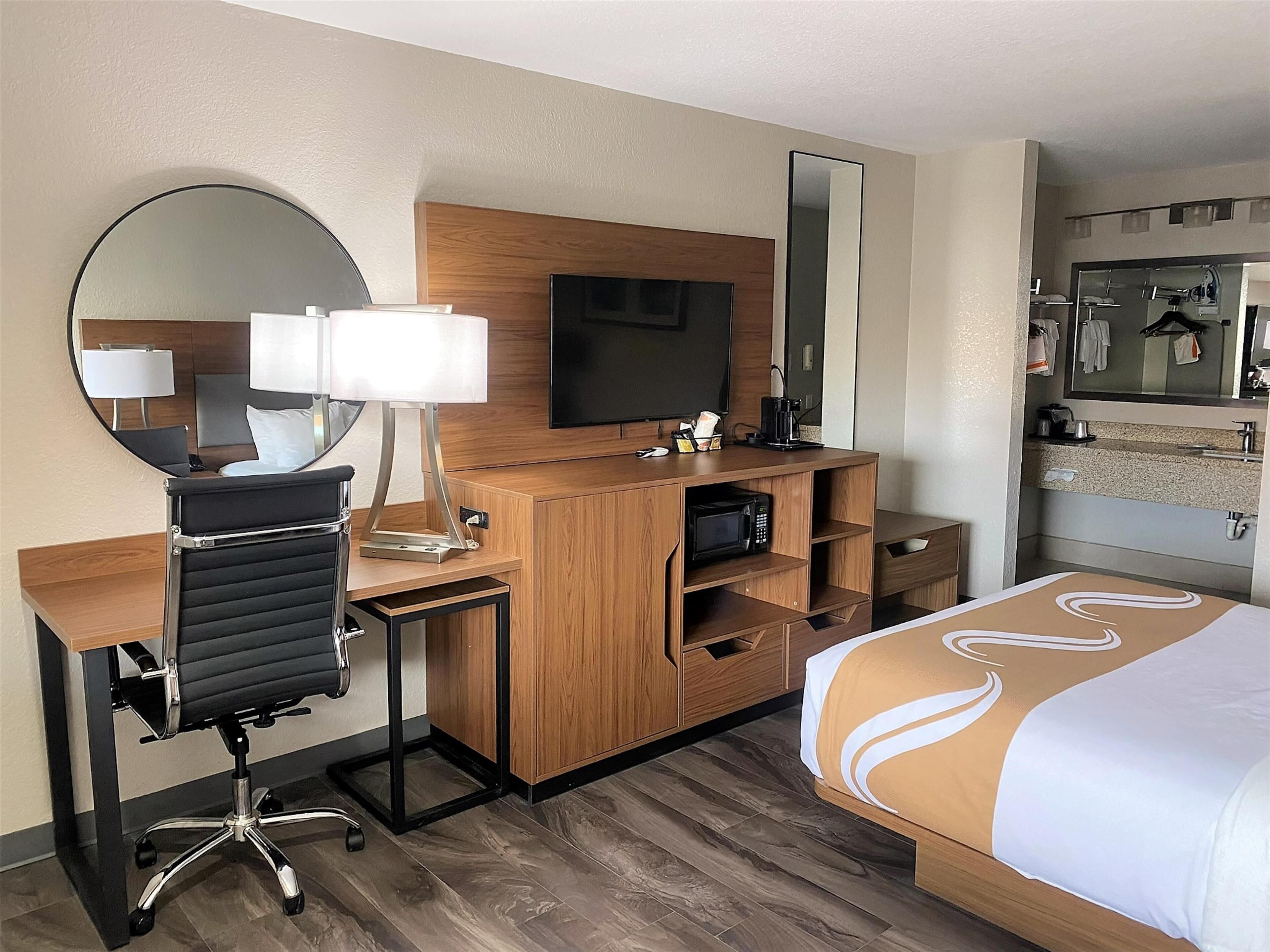 Ein Hotelzimmer mit einem Holztisch, einem schwarzen Bürostuhl, einem Fernseher, einem Spiegel, einem Bett mit weißer und brauner Bettdecke sowie einem Badezimmer mit Waschbecken und Spiegel.