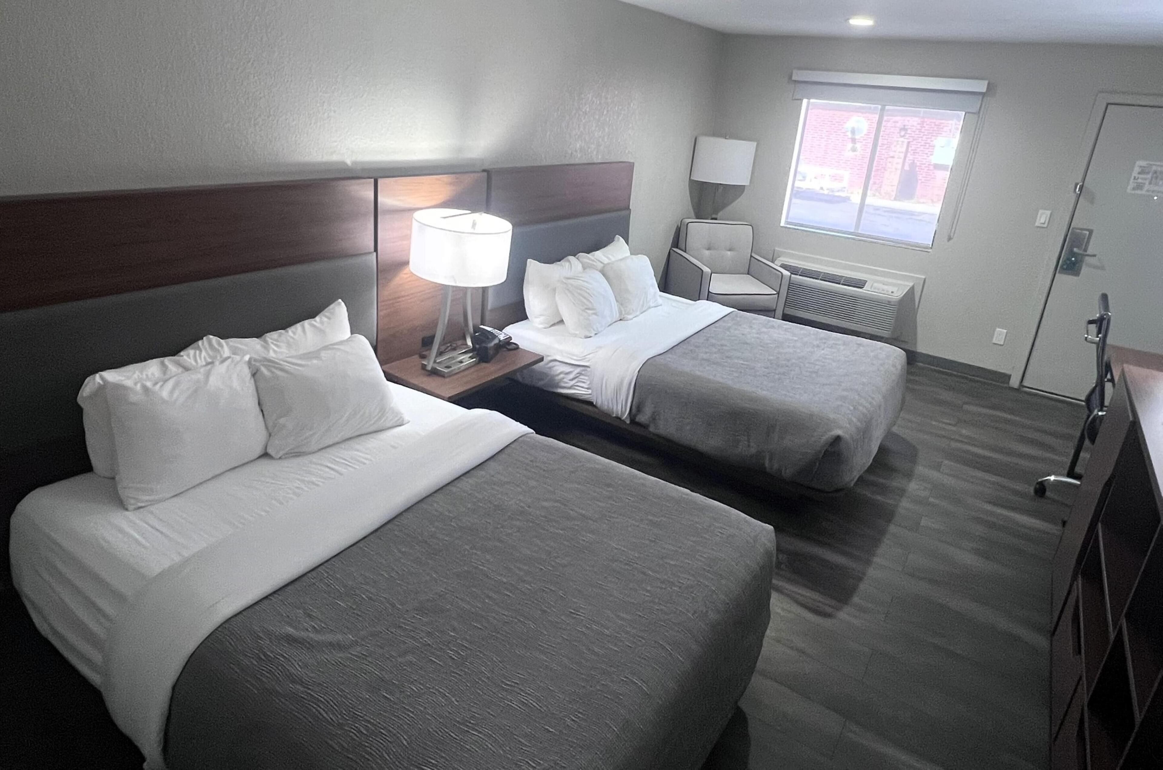 A hotel room with two beds, a nightstand with a lamp, a chair, and a window with blinds.