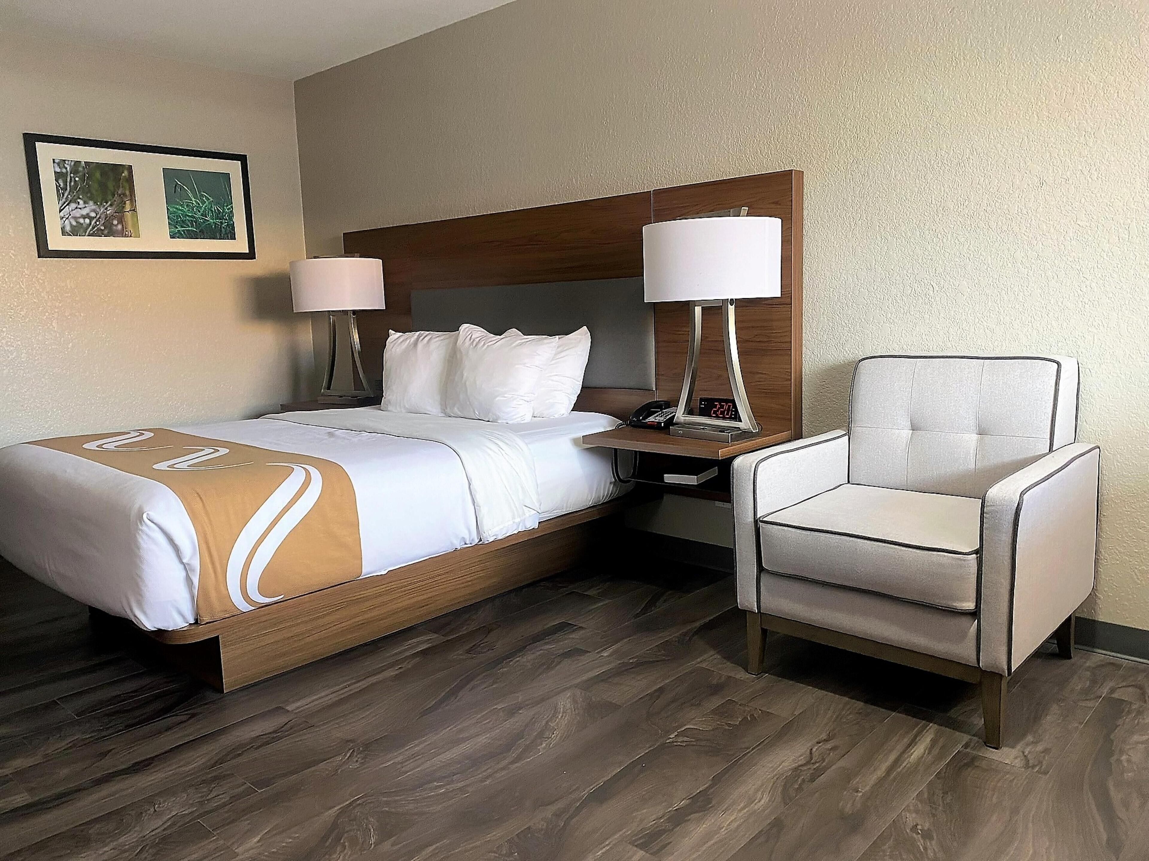 Una habitación de hotel con una cama de madera, ropa de cama blanca, dos lámparas de noche, una mesita de noche y una silla.