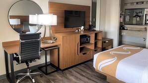 Hotellihuone, jossa on puinen työpöytä, musta toimistotuoli, televisio, peili, valko-ruskea päiväpeiteellä varustettu sänky sekä kylpyhuone, jossa on pesuallas ja peili.