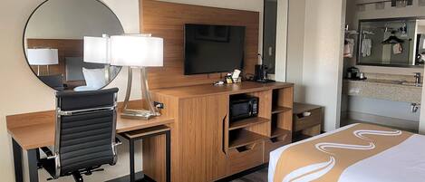 Habitación de hotel con un escritorio de madera, una silla de oficina negra, un televisor, un espejo y una cama con cubrecama blanco y marrón, además de un baño con lavamanos y espejo.