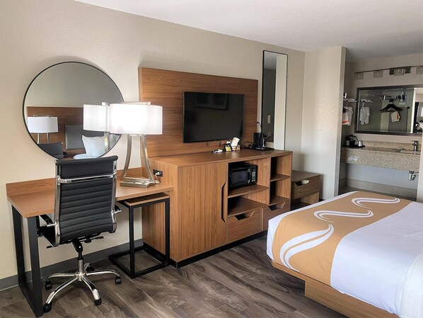 A hotel room with a wooden desk, a black office chair, a television, a mirror, a bed with a white and brown bedspread, and a bathroom with a sink and mirror.