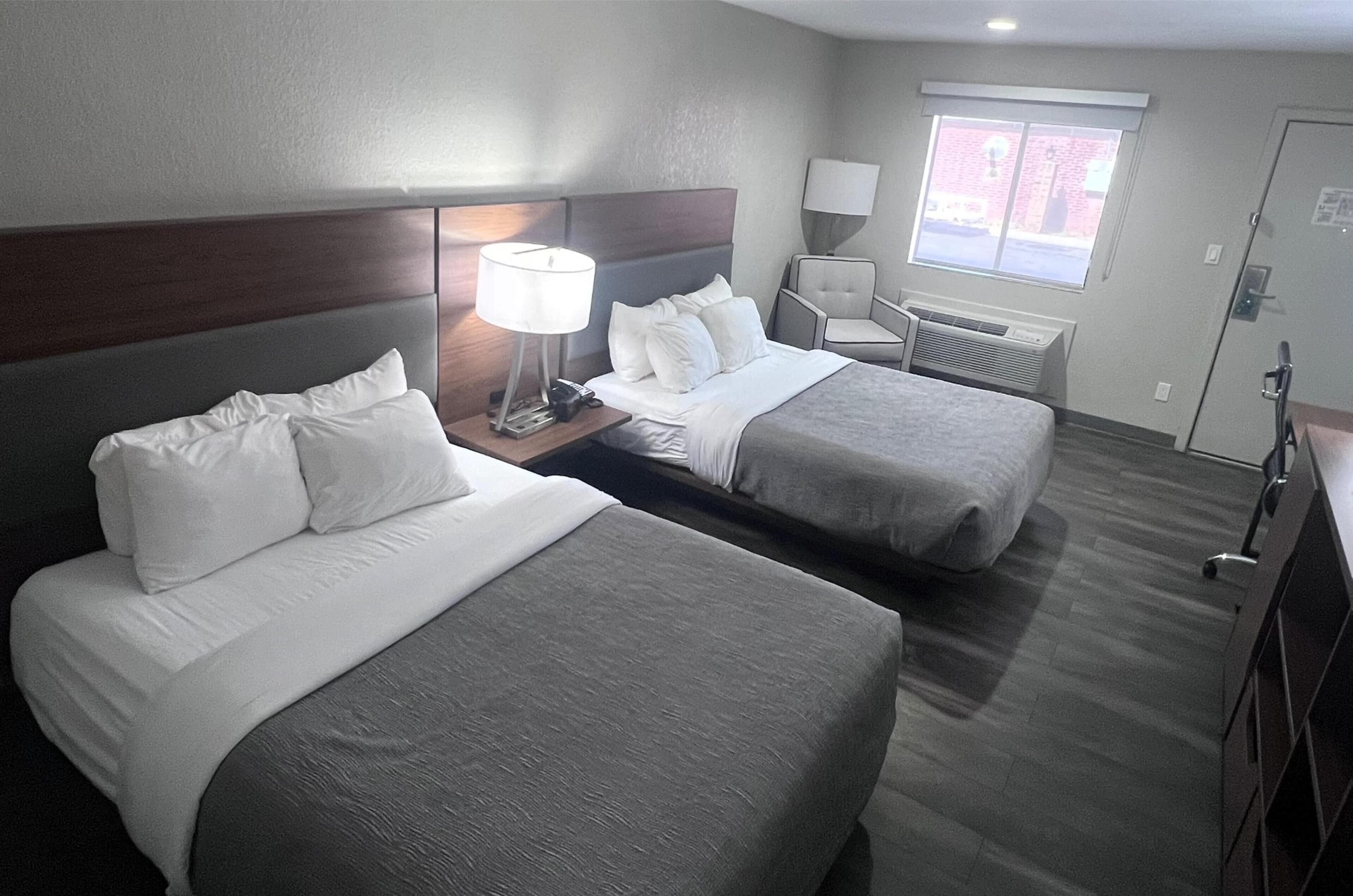 A hotel room with two beds, a nightstand with a lamp, a chair, and a window with blinds.