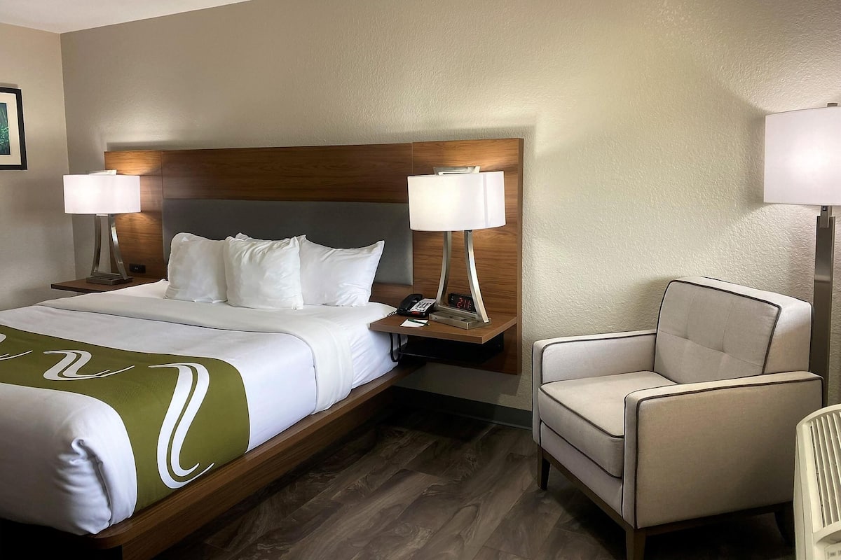 Ein Hotelzimmer mit einem Bett, zwei Nachttischlampen, einem Sessel und einem Nachttisch mit Telefon und digitaler Uhr.