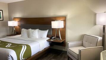 Una habitación de hotel con una cama, dos lámparas de noche, una silla y una mesita de noche con un teléfono y un reloj digital.