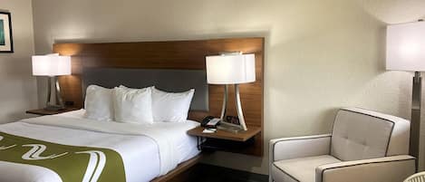 Una habitación de hotel con una cama, dos lámparas de noche, una silla y una mesita de noche con un teléfono y un reloj digital.