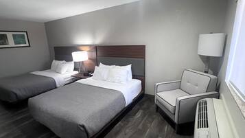 A hotel room with two beds, a chair, a desk with a lamp, and a heater.