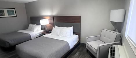A hotel room with two beds, a chair, a desk with a lamp, and a heater.