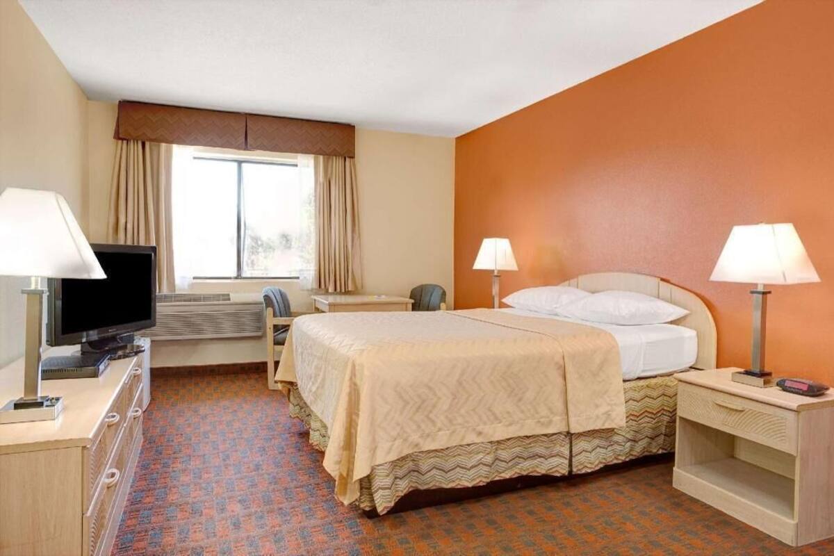 A hotel room with a bed, two bedside lamps, a television, a desk, and a window with curtains.