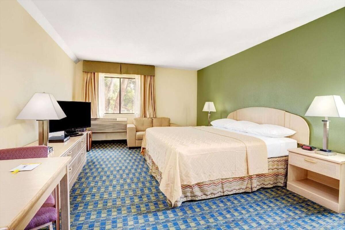 A hotel room with a bed, a desk with a television, a chair, a lamp, and a window with curtains.