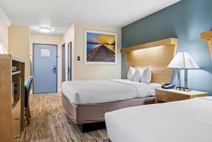 Habitación de hotel con dos camas, un escritorio, una silla, una mesita de noche con lámpara y un cuadro de atardecer en la pared.
