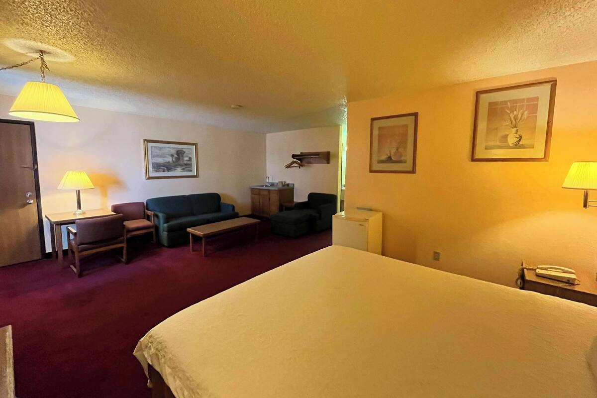 A hotel room with a bed, a desk with a lamp, a sofa, a small table, and a refrigerator.