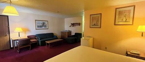 A hotel room with a bed, a desk with a lamp, a sofa, a small table, and a refrigerator.