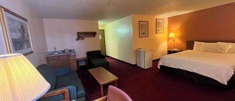A hotel room with a bed, a desk, a chair, a small table, and a lamp.