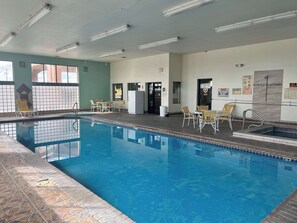An indoor swimming pool with clear water, surrounded by tiled walls and a tiled floor. There are several lounge chairs and tables around the pool area.