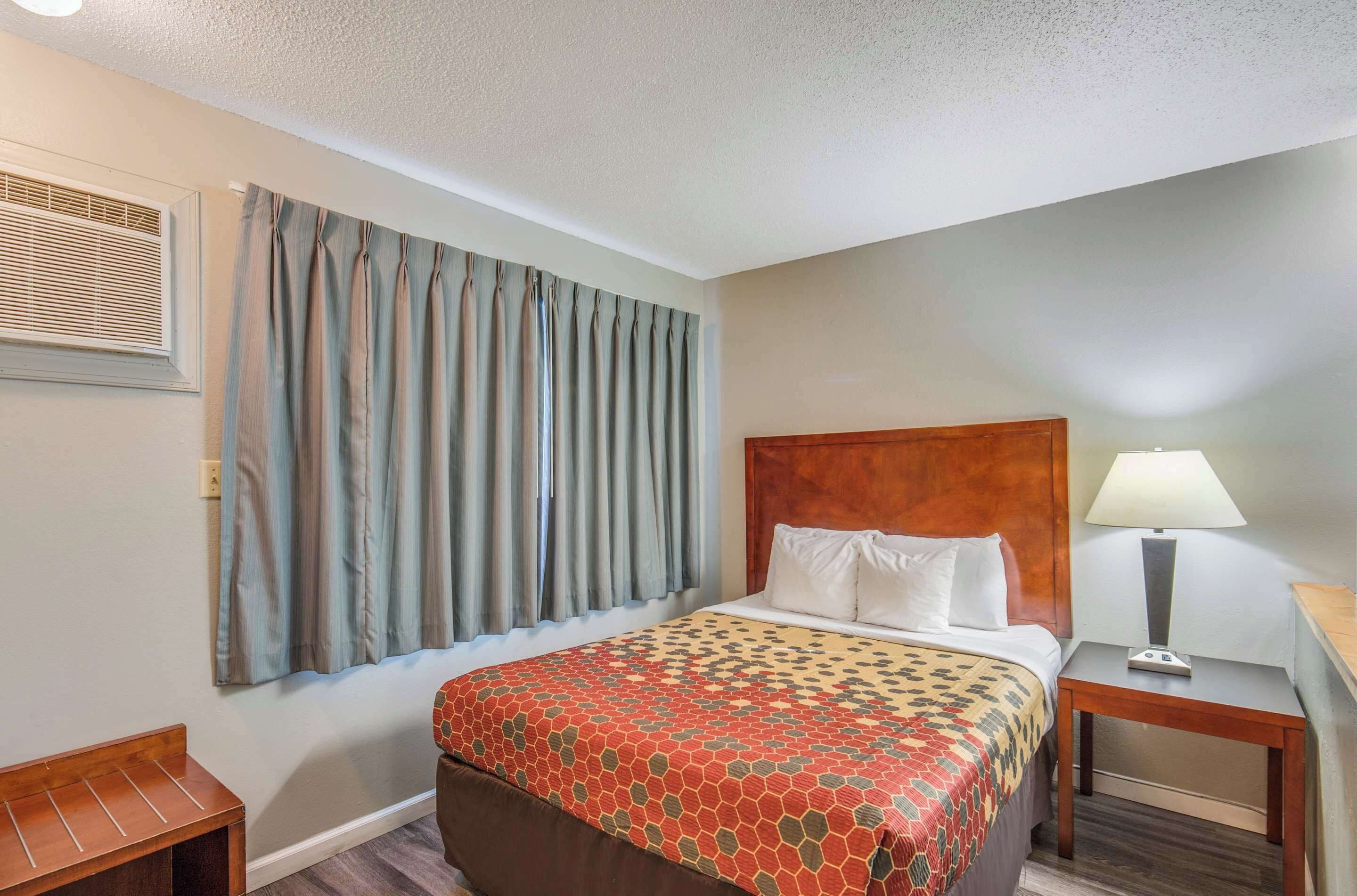 A hotel room with a bed, a nightstand with a lamp, a bench, and a window with curtains.