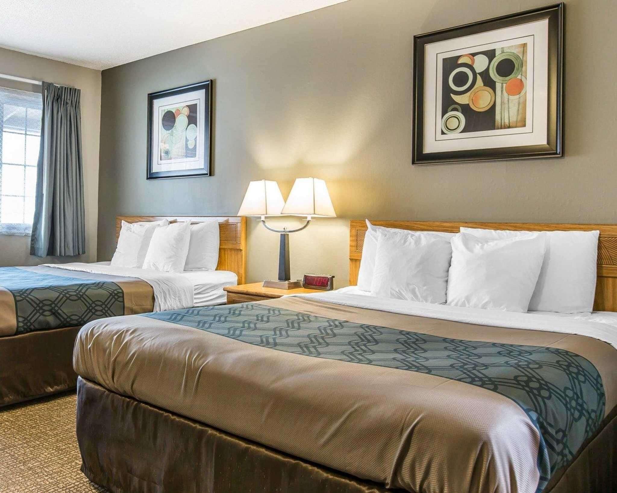 A hotel room with two beds, a nightstand with a lamp, and two framed artworks on the wall.