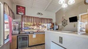 A small, clean kitchen area with a refrigerator, a microwave, and a coffee machine. There is a window with curtains, a small sink, and a countertop with a coffee maker and a toaster.