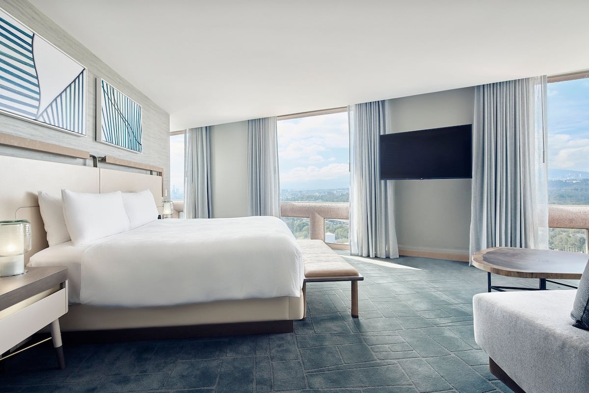 Een moderne hotelkamer met een groot bed, nachtkastje, televisie, balkon met gordijnen en uitzicht op de stad.