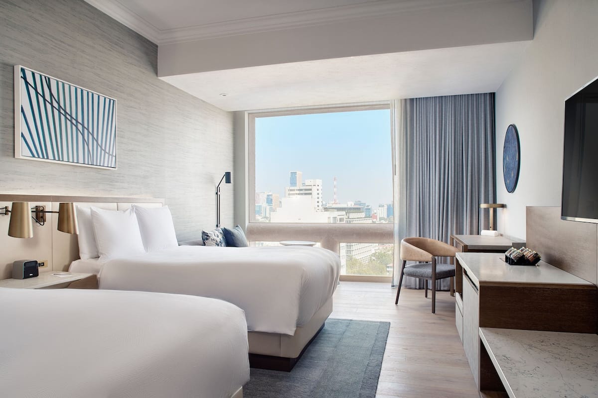 Een moderne hotelkamer met twee bedden, een bureau en een groot raam met uitzicht op de stad.