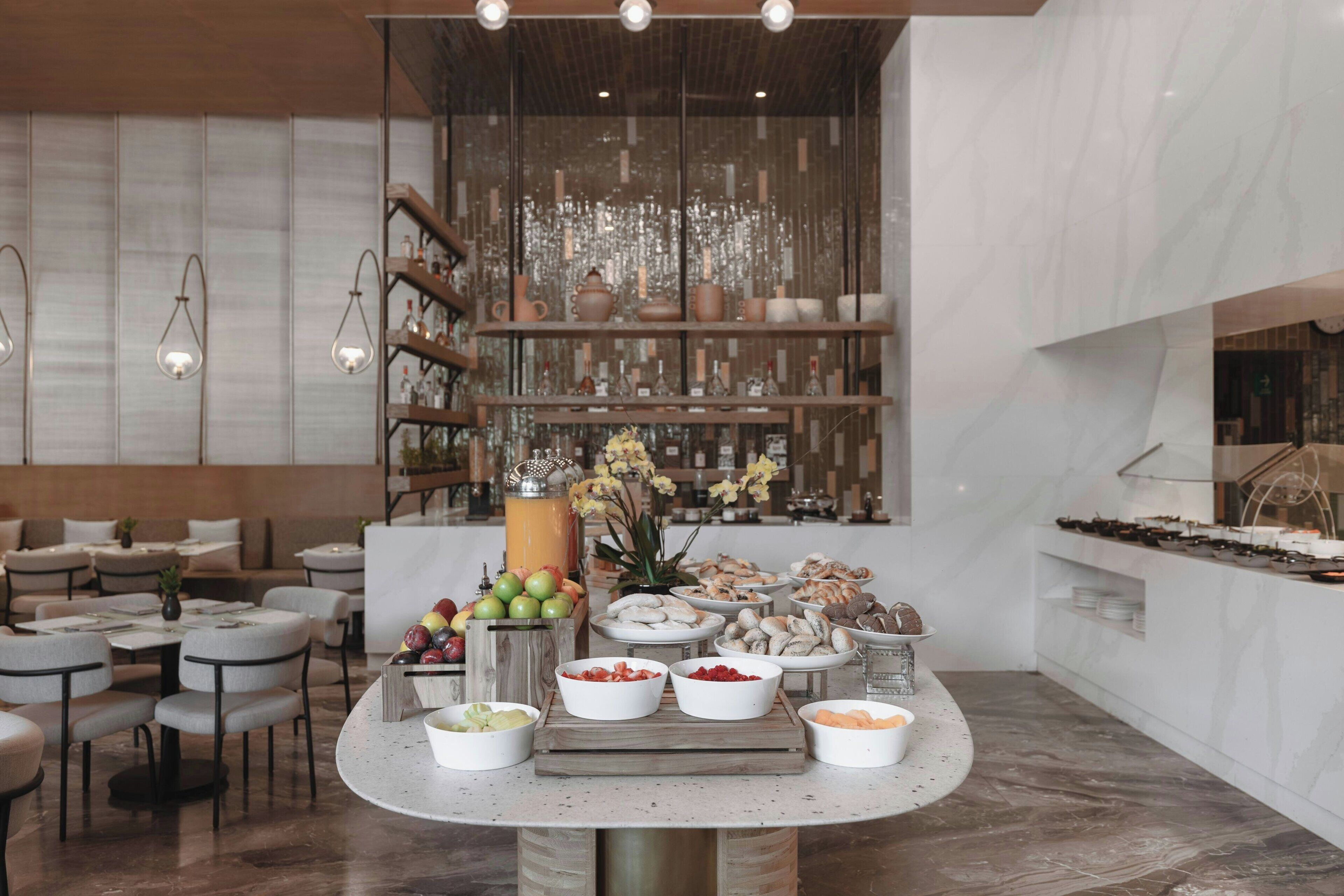 Un comedor moderno con mesas redondas, un bufé con diferentes opciones de comida y una pared decorativa con luces colgantes.