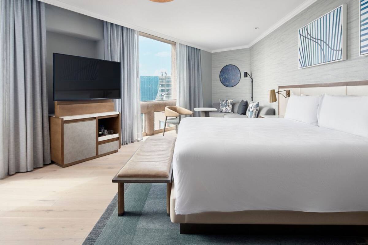 Een moderne hotelkamer met een groot bed, een flatscreen televisie, een zithoek met een tafel en stoelen, en een balkon met uitzicht over de stad.