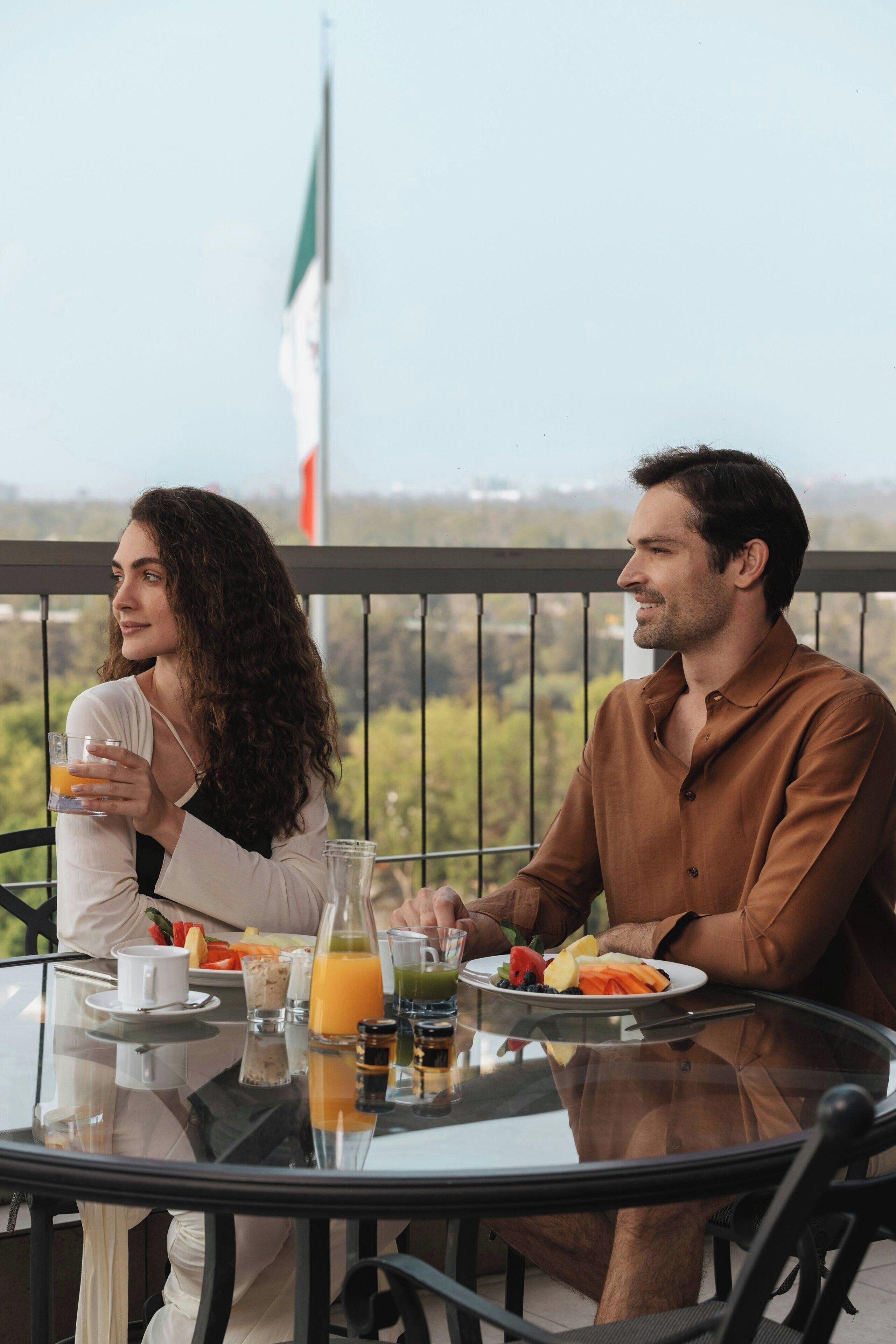 A couple dining at an outdoor table with a city view, a flag, and a glass of orange juice.